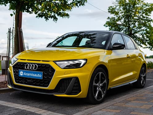 Une Audi A1 Sportback jaune reprise par Aramisauto est garée sur une place de parking en bord de route.