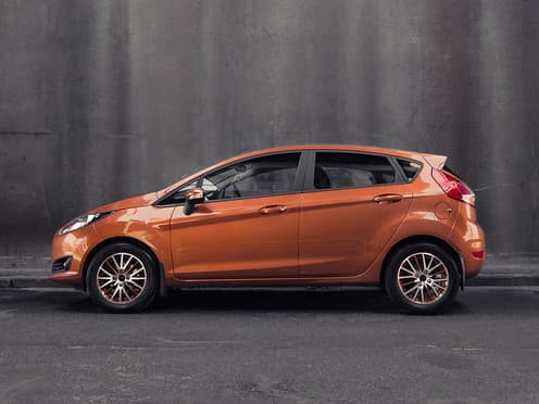 Une Ford Fiesta orange reprise par Aramisauto est à l'arrêt devant un mur en béton.